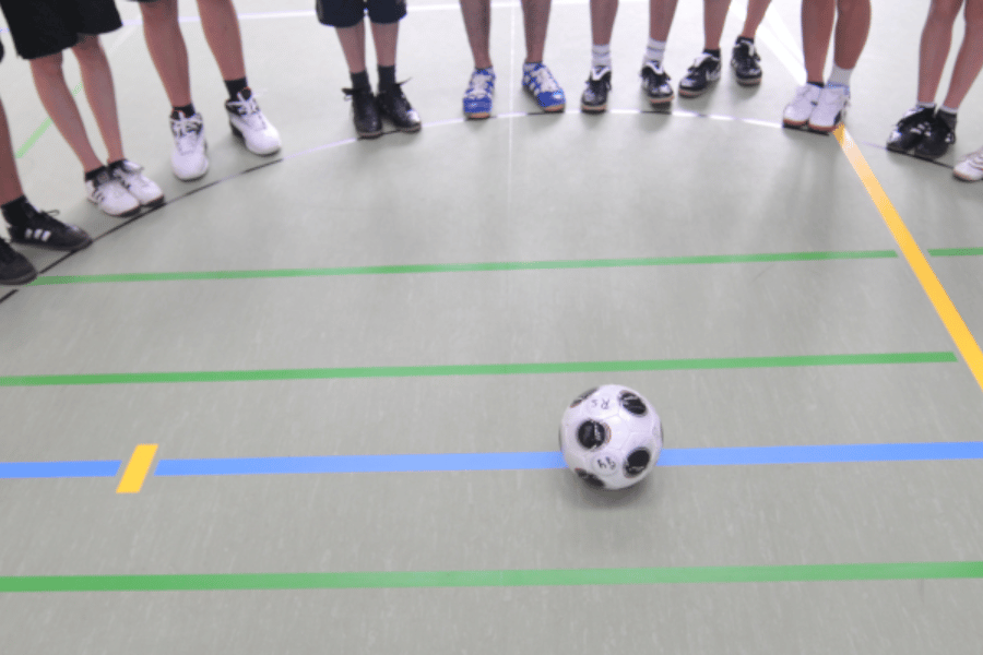Kinder stehen in einer Sporthalle um einen Fußball herum. Per Klick gelangen Sie zur Seite Hinweise für den Schulsport