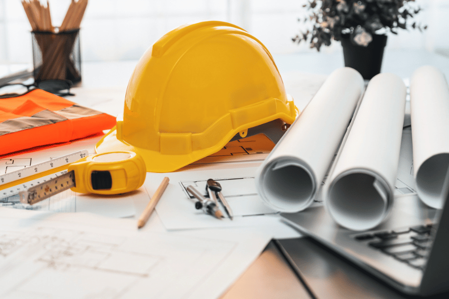 Per Klick gelangen Sie zur Seite Bau und Ausstattung. Schreibtisch mit Laptop Bauhelm und Planrollen