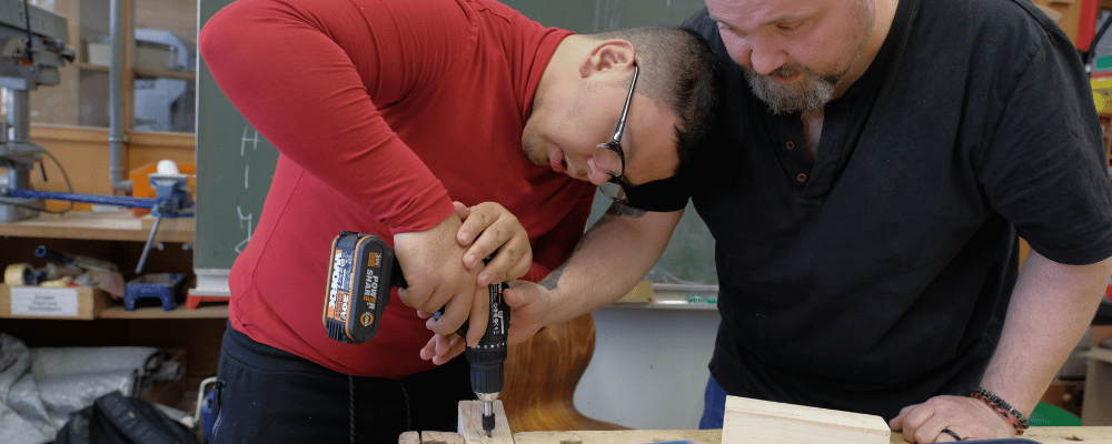 Lehrkraft gibt einem Schüler Hilfestellung beim Bohren mit einem Akkuschrauber