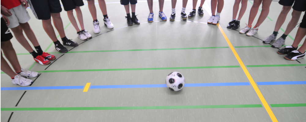 Foto: Auf dem Boden einer Sporthalle liegt ein Fußball. Im Halbkreis um den Ball stehen Schülerinnen und Schüler, deren Beine zu sehen sind.